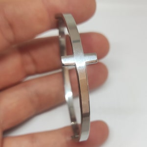 Puede incluir: Un brazalete de plata con un diseño de cruz. El brazalete está hecho de metal y tiene un acabado pulido.