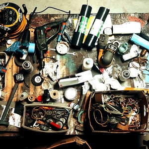 May include: A cluttered workbench with various tools and supplies, including power tools, sandpaper, wire, and a small toolbox. The workbench is made of wood and has a worn surface.