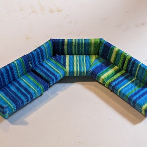 May include: A miniature sectional sofa with a vibrant blue, green, and white striped pattern. The small sofa is designed in a corner configuration, perfect for a dollhouse or miniature setting. The fabric has a textured appearance.