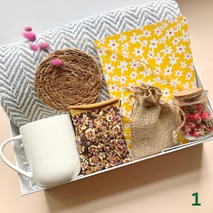 May include: A gift box filled with a white mug, a gray and white patterned blanket, a yellow floral notebook, a woven coaster, a small burlap bag, a jar of dried flowers, and a floral embroidered pouch. The box is white and contains pink dried flowers.