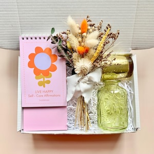 Könnte beinhalten: Geschenkbox mit einem pinkfarbenen Spiralnotizbuch mit Blumengrafik und dem Text "LIVE HAPPY Self-Care Affirmations". Enthält auch einen Trockenblumenstrauß, eine grüne Glasflasche und einen goldfarbenen Kerzenhalter.