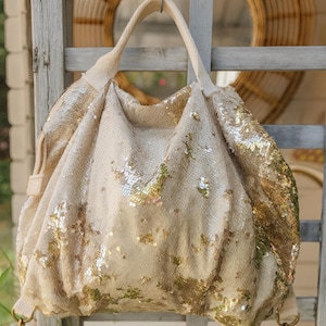 Può includere: Una grande borsa tote con paillettes color crema con tracolla e manico superiore. La borsa è impreziosita da paillettes dorate, creando un effetto scintillante. La borsa è appesa a una struttura in legno.