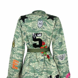 Op de afbeelding: Een groene camouflagejas met verschillende patches, waaronder een zwart nummer "5", een rode klaproos, een zwart-wit dierenhoofd en een zwart-wit gevleugeld logo met de letters "GG".