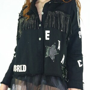 Può includere: Giacca di jeans nera con frange e borchie argentate. La giacca presenta la scritta bianca "PEACE WORLD" e una toppa a forma di stella. Un capo d'abbigliamento alla moda e audace.