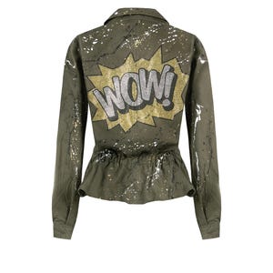 Może przedstawiać: Zielona kurtka w stylu wojskowym z białymi plamami farby i złotym i srebrnym nadrukiem "WOW!" na plecach.