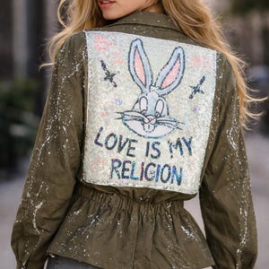 Könnte beinhalten: Olivgrüne Jacke mit weißen Farbspritzern und einem Pailletten-Design von Bugs Bunny auf dem Rücken. Das Design enthält den Text "LOVE IS MY RELIGION". Die Jacke hat eine geraffte Taille und Knöpfe.