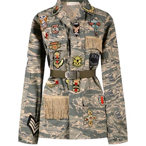 Op de afbeelding: Een camouflagejas met verschillende kleurrijke patches en een bruine riem. De jas heeft een knoopsluiting aan de voorkant en lange mouwen.