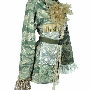 Op de afbeelding: Een groene camouflagejas met een bruine verenbies en een wit kantdetail. De jas heeft een riem en een bruin lint dat om de taille is geknoopt.