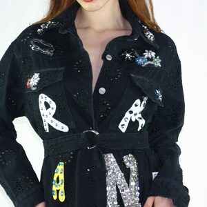 May include: Black denim jacket with decorative embellishments. The jacket features cut-out detailing, a belt, and the letters "R", "A", and "N" in white and silver. Floral and jewel accents adorn the collar and pockets.