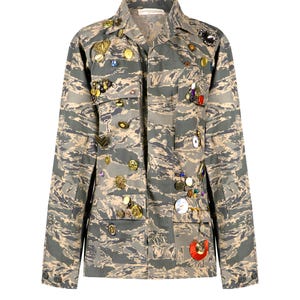 Op de afbeelding: Een camouflagejas met een verscheidenheid aan knopen, pinnen en andere versieringen. De jas heeft een lange mouw en een knoopsluiting.