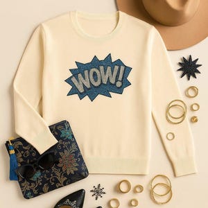 Puede incluir: Jersey color crema con un gráfico "WOW!" azul y blanco. La imagen también presenta un sombrero marrón, un clutch estampado con gafas de sol, bailarinas negras y joyas doradas, incluyendo anillos y pulseras. También hay un accesorio en forma de estrella y de copo de nieve.