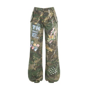 Könnte beinhalten: Camouflagehose mit weißen Farbstichen und bunten Blumen- und Grafikdesigns. Die Hose hat eine weite Beinweite und eine lockere Passform. Der Text "THE" und "LOOK" ist auf der Hose aufgedruckt.
