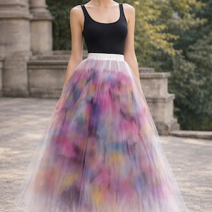 May include: A woman wearing a black tank top and a long, colorful tulle skirt. The skirt features a watercolor-like design with shades of pink, purple, blue, and yellow. The skirt has a white waistband. The woman is wearing gold sandals.