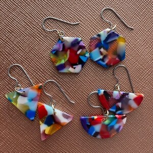 May include: A collection of colorful, geometric earrings. The earrings feature a mosaic pattern of red, blue, yellow, and white pieces. The earrings are in the shape of a hexagon, triangle, and half-circle, with silver-colored ear wires.