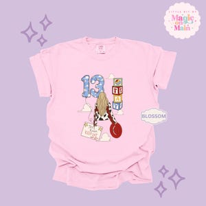 May include: A light pink t-shirt featuring a graphic of a person with long blonde hair wearing a cow-print jacket and holding a red hat. The design includes a blue cloud-patterned number 13 balloon, stacked alphabet blocks spelling TAY, and a sign reading Tay's Room Keep Out.