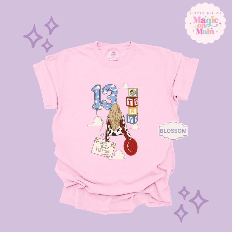 May include: A light pink t-shirt featuring a graphic of a person with long blonde hair wearing a cow-print jacket and holding a red hat. The design includes a blue cloud-patterned number 13 balloon, stacked alphabet blocks spelling TAY, and a sign reading Tay's Room Keep Out.
