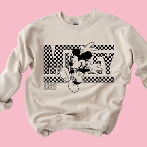 Puede incluir: Una sudadera beige con un dise&ntilde;o a cuadros en blanco y negro con Mickey Mouse y la palabra "MICKEY" con el a&ntilde;o "1928" impreso debajo.