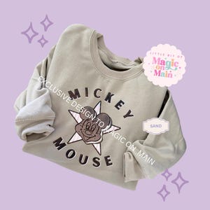 May include: Sand-colored sweatshirt featuring a Mickey Mouse design. The design includes the text "MICKEY MOUSE" and a star graphic. The sweatshirt has a crew neck and long sleeves. The text "LITTLE BIT OF MAGIC ON MAIN" is also visible.