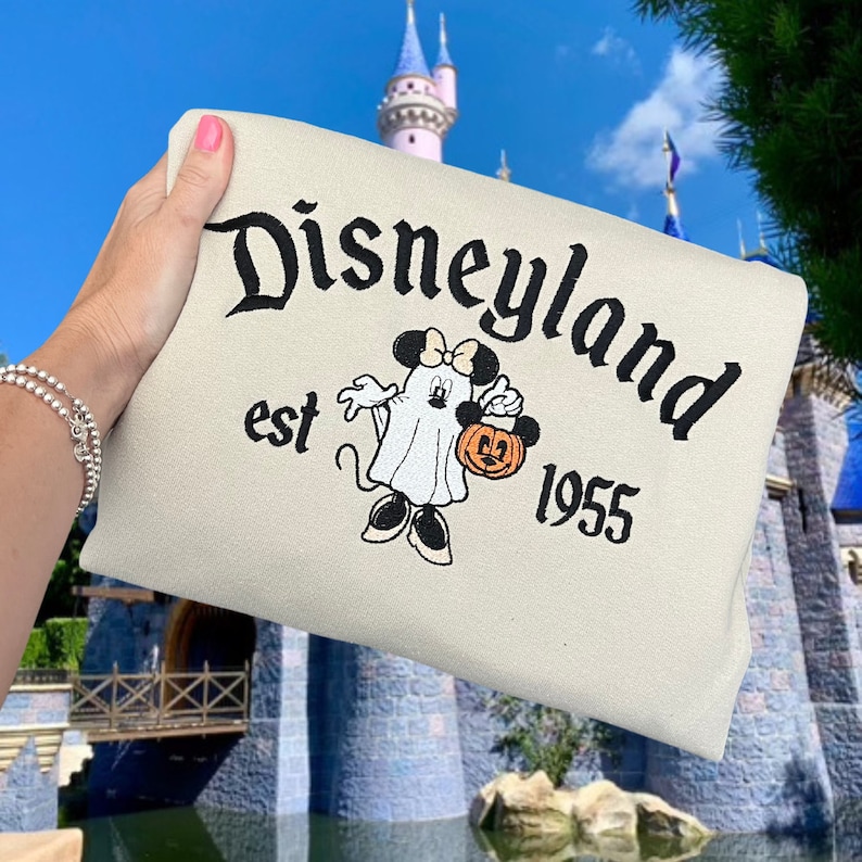 K&ouml;nnte beinhalten: Ein beiger Pullover mit einem schwarzen gestickten Design von Mickey Mouse, der als Geist verkleidet ist und einen K&uuml;rbis h&auml;lt. Der Text "Disneyland est 1955" ist ebenfalls auf dem Pullover gestickt.