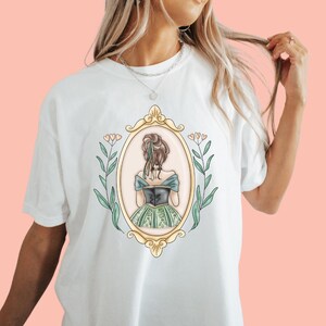 Puede incluir: Camiseta blanca con un gr&aacute;fico de una mujer con un vestido verde y dorado, de pie en un marco ovalado dorado con detalles florales.