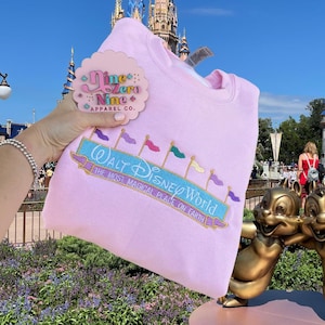 May include: Pink sweatshirt featuring a Walt Disney World logo and a Nine Zero Nine Apparel Co. pin. The logo has a blue banner with white text and colourful flags.