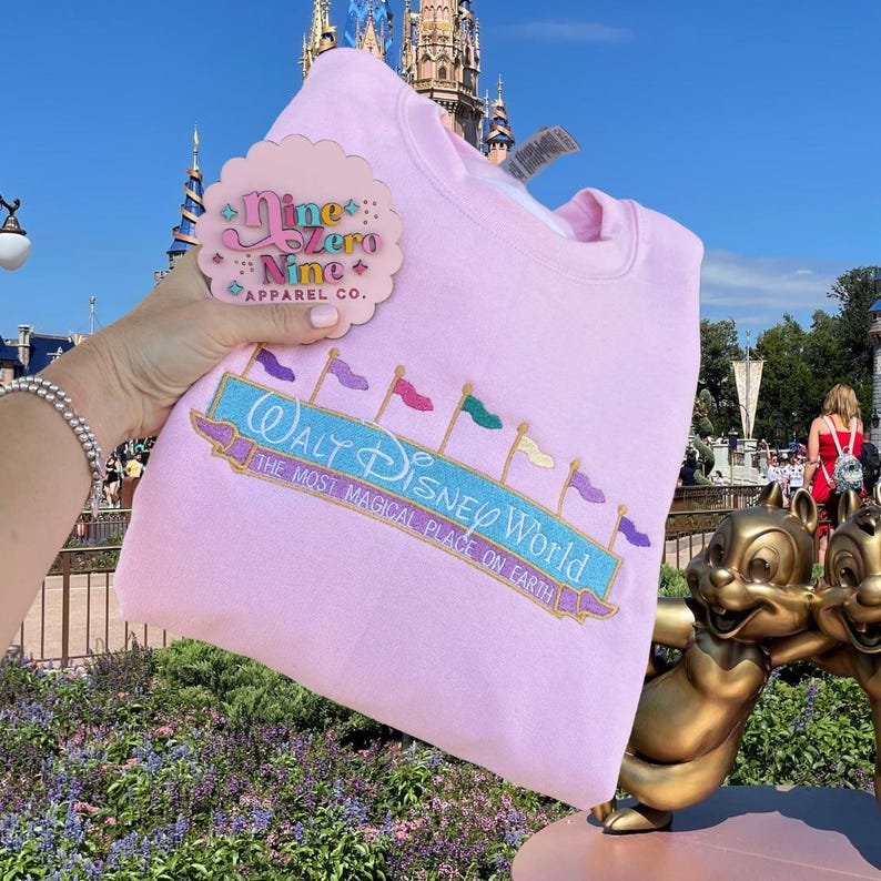May include: Pink sweatshirt featuring a Walt Disney World logo and a Nine Zero Nine Apparel Co. pin. The logo has a blue banner with white text and colourful flags.