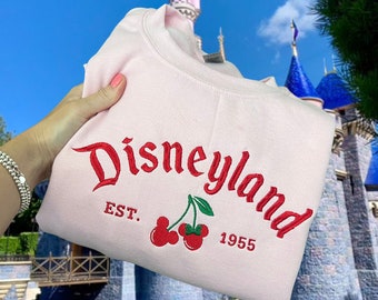 Może przedstawiać: Różowa bluza z haftowanym czerwonym tekstem "Disneyland Est. 1955" z dwoma czerwonymi wiśniami i zielonymi szypułkami.