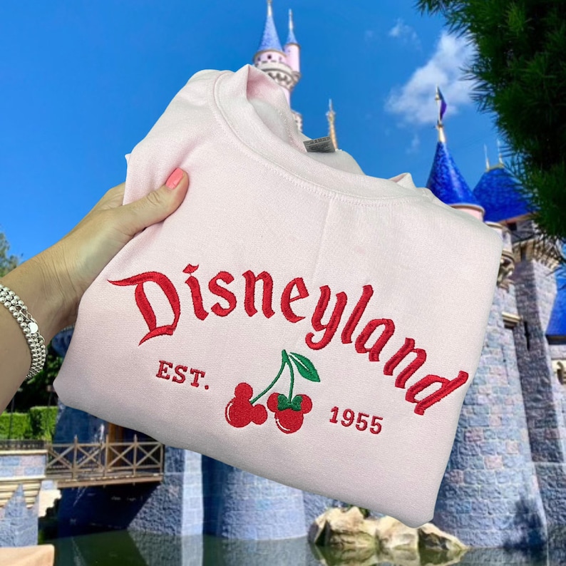 May include: Pink sweatshirt with red embroidered text that reads "Disneyland Est. 1955" with two red cherries and green stems.