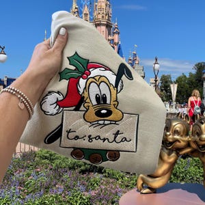 May include: A beige tote bag with an embroidered image of Pluto dressed as Santa Claus. The image includes a red Santa hat, green holly, and a sign that says "to: santa".