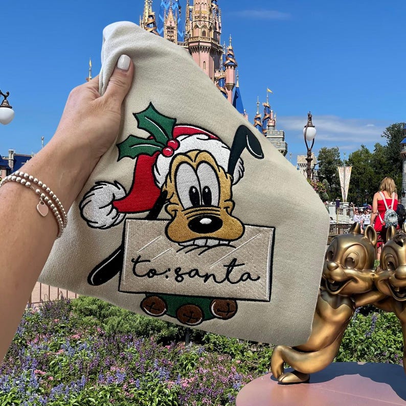 May include: A beige tote bag with an embroidered image of Pluto dressed as Santa Claus. The image includes a red Santa hat, green holly, and a sign that says "to: santa".