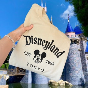 Puede incluir: Una sudadera beige con un gr&aacute;fico de Mickey Mouse negro y el texto "Disneyland EST 1983 TOKYO".
