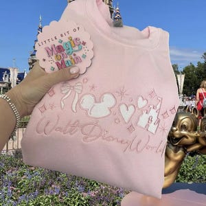 May include: A light pink sweatshirt with white embroidered Disney-themed designs, including Mickey Mouse ears, hearts, and a castle. The text "Walt Disney World" is embroidered below the designs. A tag with the text "Little Bit of Magic on Main" is attached.