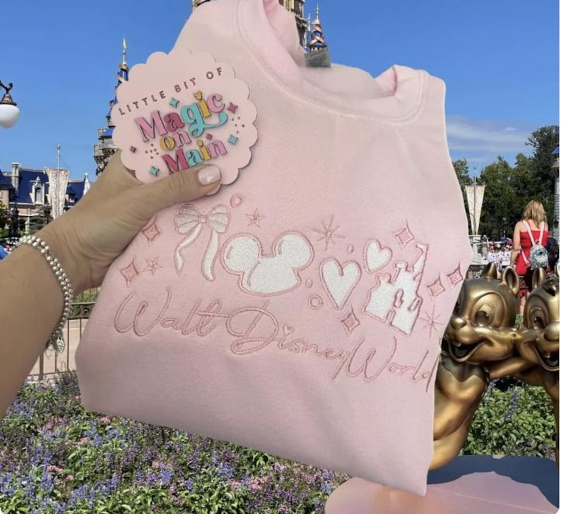 May include: A light pink sweatshirt with white embroidered Disney-themed designs, including Mickey Mouse ears, hearts, and a castle. The text "Walt Disney World" is embroidered below the designs. A tag with the text "Little Bit of Magic on Main" is attached.