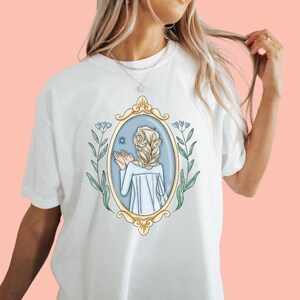 May include: White t-shirt with a graphic of a woman with blonde hair in a blue and gold frame. The woman is reaching out to a small white star. The frame is surrounded by green leaves and blue flowers.