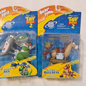 May include: Two Toy Story 2 action figures in their packaging. The figure on the left is Rex, a green dinosaur with a jetpack. The figure on the right is Bullseye, a brown horse with a saddle and a rocket launcher. Both figures are part of the Rocket Force line.