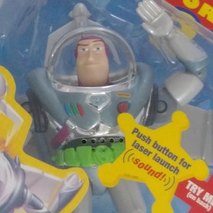 May include: A silver and green Buzz Lightyear action figure from the Toy Story franchise. The figure has a push button on the chest that activates a laser launch sound effect. The figure is wearing a space suit with a helmet and has a green belt with a silver buckle.