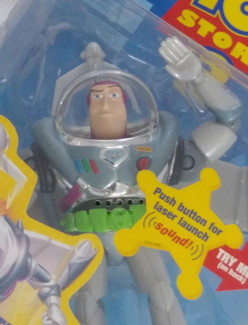 May include: A silver and green Buzz Lightyear action figure from the Toy Story franchise. The figure has a push button on the chest that activates a laser launch sound effect. The figure is wearing a space suit with a helmet and has a green belt with a silver buckle.