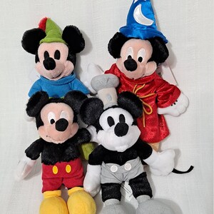 Op de afbeelding: Vier pluche Mickey Mouse-speelgoed in verschillende outfits. E&eacute;n Mickey draagt een rode robe en een blauwe tovenaarshoed met een halve maan en een ster. Een andere Mickey draagt een blauw shirt en een groene hoed met een veer. Een derde Mickey draagt een rode broek en een zwart shirt. De vierde Mickey draagt een grijze broek en een zwart shirt.