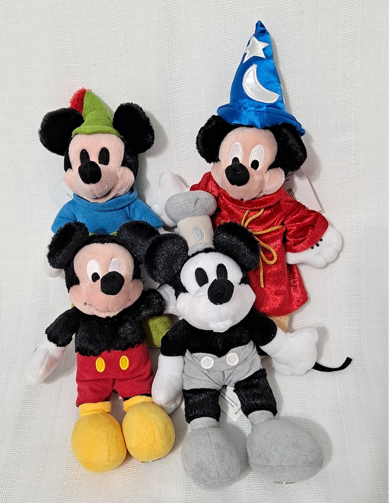 Op de afbeelding: Vier pluche Mickey Mouse-speelgoed in verschillende outfits. E&eacute;n Mickey draagt een rode robe en een blauwe tovenaarshoed met een halve maan en een ster. Een andere Mickey draagt een blauw shirt en een groene hoed met een veer. Een derde Mickey draagt een rode broek en een zwart shirt. De vierde Mickey draagt een grijze broek en een zwart shirt.