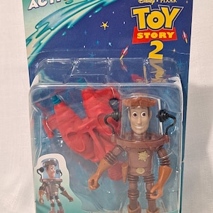 Pode incluir: Uma figura de a&ccedil;&atilde;o de pl&aacute;stico do Woody de Toy Story 2, vestindo um traje espacial marrom e segurando um esguicho de &aacute;gua vermelho. A embalagem &eacute; azul e apresenta o logotipo do Toy Story 2.