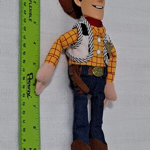 May include: A plush toy of Woody from the Toy Story franchise. Woody is wearing his signature brown cowboy hat, a yellow and red plaid shirt, a brown vest with a white and black striped pattern, blue jeans, and brown cowboy boots. He is holding a brown holster with a brown gun.