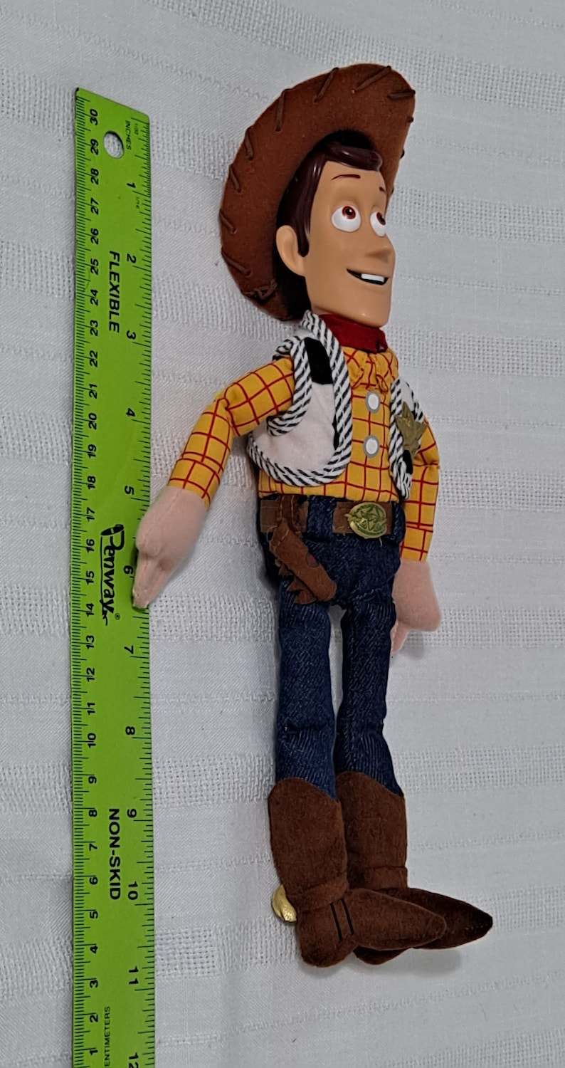 May include: A plush toy of Woody from the Toy Story franchise. Woody is wearing his signature brown cowboy hat, a yellow and red plaid shirt, a brown vest with a white and black striped pattern, blue jeans, and brown cowboy boots. He is holding a brown holster with a brown gun.