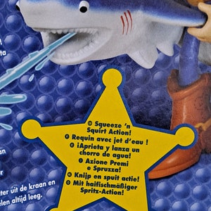 May include: A yellow star with a blue shark on top. The star has text in multiple languages that says "Squeeze 'n Squirt Action!"  The shark has a blue body with white teeth and a red stripe on its back. The shark is holding a blue water squirter in its mouth.