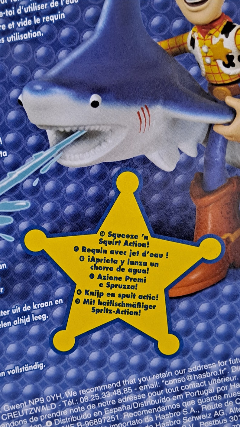 May include: A yellow star with a blue shark on top. The star has text in multiple languages that says "Squeeze 'n Squirt Action!"  The shark has a blue body with white teeth and a red stripe on its back. The shark is holding a blue water squirter in its mouth.