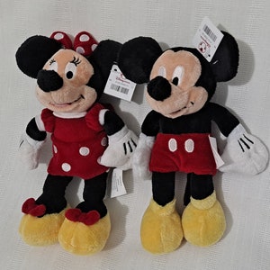 May include: Two plush toys, Mickey Mouse and Minnie Mouse, are standing side-by-side. Mickey Mouse is wearing red shorts and black shoes. Minnie Mouse is wearing a red dress with white polka dots and black shoes. Both toys have black eyes and white gloves.