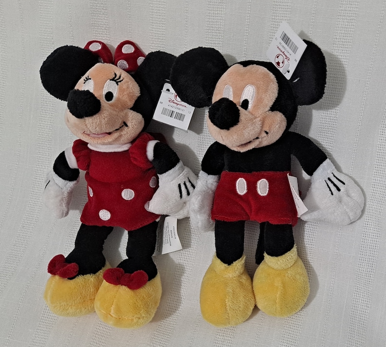 May include: Two plush toys, Mickey Mouse and Minnie Mouse, are standing side-by-side. Mickey Mouse is wearing red shorts and black shoes. Minnie Mouse is wearing a red dress with white polka dots and black shoes. Both toys have black eyes and white gloves.
