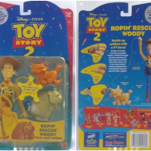 May include: Toy Story 2 action figure of Woody from the movie. Woody is wearing his classic cowboy outfit and holding a rope. He is standing next to a brown and white dog figure. The packaging is blue and yellow with the Toy Story 2 logo and the words "Ropin' Rescue Woody".