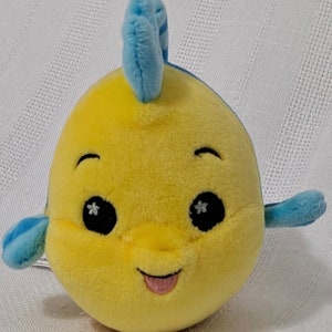 May include: Yellow plush toy shaped like a fish with blue fins and a blue fin on top of its head. The fish has black eyes with white stars, a pink tongue sticking out, and a smiling face.
