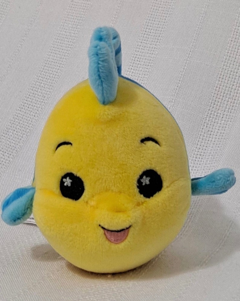 May include: Yellow plush toy shaped like a fish with blue fins and a blue fin on top of its head. The fish has black eyes with white stars, a pink tongue sticking out, and a smiling face.