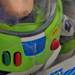 May include: A green and white Buzz Lightyear toy with a blue space ranger emblem on the chest. The toy is in a clear plastic package with the words "ORIGINAL - EL ORIGINAL" and "Buzz" printed on the bottom. The toy has a red button on the side and a yellow label that says "LIGHTYEAR".